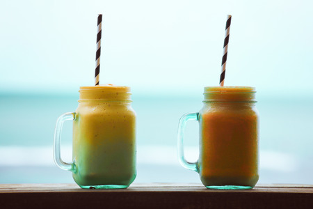 Selective focus on glass of mango and papaya smoothie on wooden table with blue oceans background. Healthy summer drink concept.の写真素材