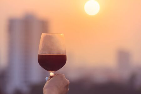 Womanâs hand holding red wine glasses on balcony during sunset, celebration conceptの写真素材