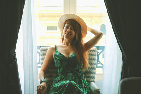 Smiling asian woman with hat sitting on chair, feeling happy and positiveの写真素材