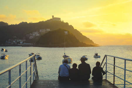 Selective focus on mountain and boats. Happy family sitting on staire and watching the sunset in San Sebastian, Spainの写真素材