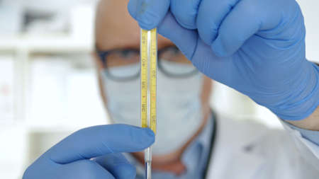 Doctor with Protection Gloves and Face Mask Keeping in His Hand a Medical Thermometer Used for Checking Human Body Temperatureの写真素材
