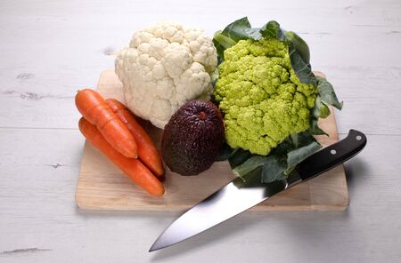 Broccoli, cauliflower, avocado and carrots with knife, on a wooden board, on white wooden tableの写真素材