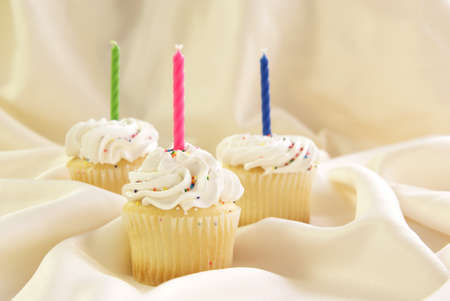 Three cupcakes with candles and colorful candy sprinkles in whipped cream icing.の写真素材