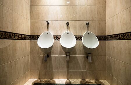 Three bright white urinals fitted to wall covered with light brown stone tiles in hotel. Low angle front view shotの写真素材