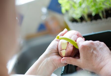 Senior woman peeling red potatoes, preparing food. Horizontal orientation with selective focus on potatoe and peeler. Natural day lightの写真素材