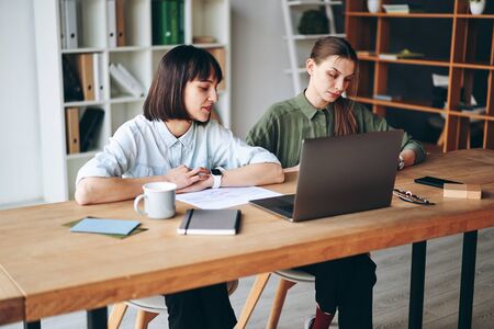 Two young woman working, looking at laptop screen, making notes and drinking coffeeの写真素材