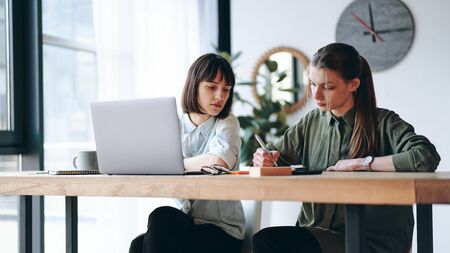Two young woman working in modern office using laptop and smart phone, making notesの写真素材