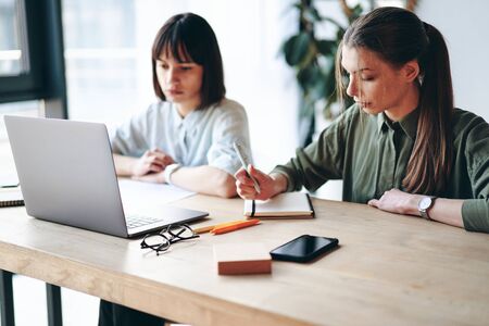 Two young woman working in modern office using laptop and smart phone, making notesの写真素材