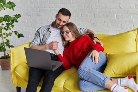 Young cute couple looking at laptop screen siting on the yellow couchの写真素材
