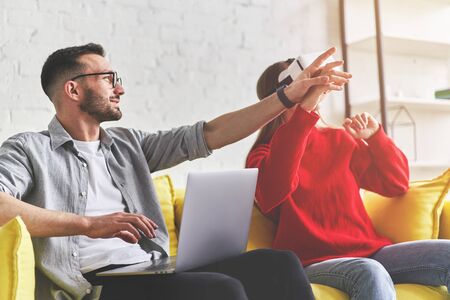 Young smiling couple in VR glasses playing video game with laptop on yellow couchの写真素材