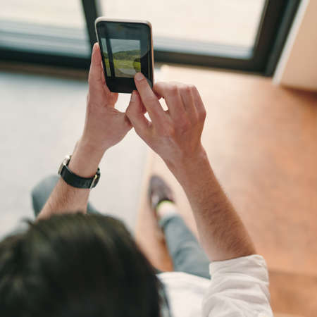 Close up of young hipster guy taking photo on his mobile phone camera of a beautiful landscape through the windowの写真素材