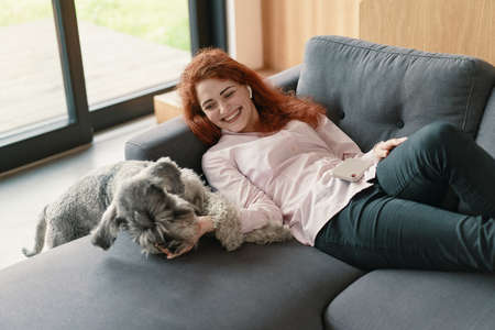 Beautiful redhead woman resting on a sofa laughing and cuddling a dogの写真素材