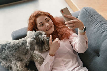 Girl with red hair and charming smile lying on sofa and making selfie with her pet