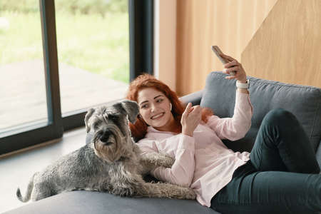 Portrait of beautiful young woman with her dog using mobile phone at homeの写真素材