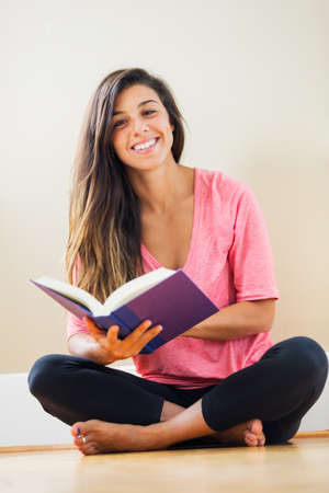 Portrait of beautiful happy young woman sitting on floor reading a book smiling - Indoor