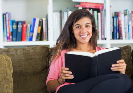 beautiful happy young woman reading a book sitting on a sofa in a living room