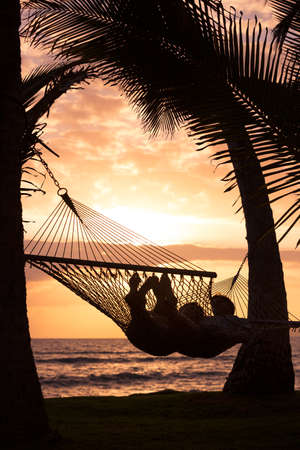 Silhouette of Romantic couple relaxing in tropical hammock at sunsetの写真素材