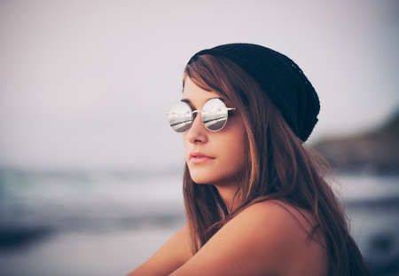 Fashion portrait of young hipster woman with hat and sunglasses on the beach at sunset, retro style color tonesの写真素材