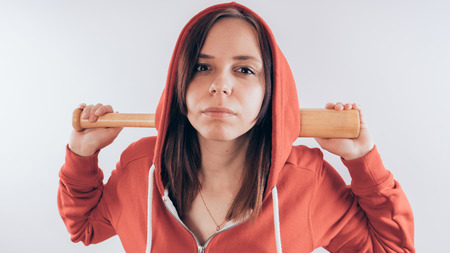 Girl athlete with a baseball bat in a red hoodie. Girl or criminal woman holding baseball bat in hand on white background. Body art. Sport and trainingの写真素材