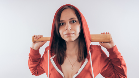 Girl athlete with a baseball bat in a red hoodie. Girl or criminal woman holding baseball bat in hand on white background. Body art. Sport and trainingの写真素材