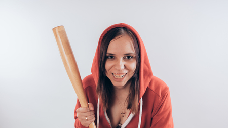 Girl athlete with a baseball bat in a red hoodie. Girl or criminal woman holding baseball bat in hand on white background. Body art. Sport and trainingの写真素材