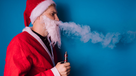 Guy dressed as Santa Claus vaping electronic cigarette closeup. Man smoking e-cigarette to quit tobacco. Vapor and alternative nicotine free smoking concept, copy spaceの写真素材