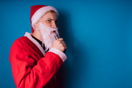 Guy dressed as Santa Claus vaping electronic cigarette closeup. Man smoking e-cigarette to quit tobacco. Vapor and alternative nicotine free smoking concept, copy spaceの写真素材
