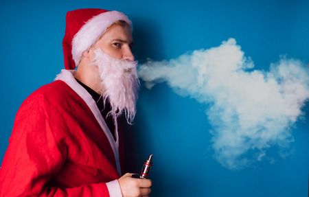 Guy dressed as Santa Claus vaping electronic cigarette closeup. Man smoking e-cigarette to quit tobacco. Vapor and alternative nicotine free smoking concept, copy spaceの写真素材