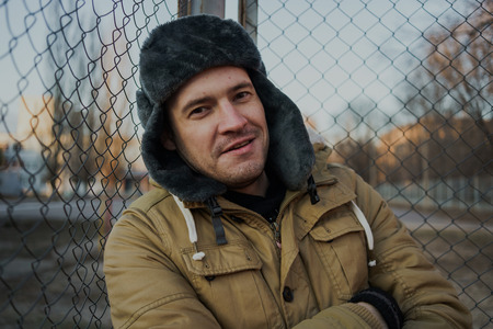 Happy handsome young man in cap with earflaps. The young man in the fur hat. standing on a cloudy day. emotional portrait of a student. street styleの写真素材