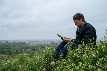 Side view of young handsome man in glasses and warm jacket reading book in open space on background...