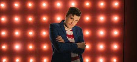 Young male standing against illuminated wall. Trendy man looking at camera on red background with...