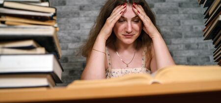 Tired female student reading among books. Pensive young woman sitting at table with pile of book and...