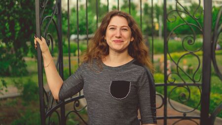 Smiling woman against fence in garden. Content woman looking dreamily standing in green park against ornamental iron fenceの写真素材