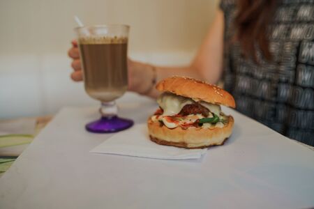 Food photography. Hot cappuccino with hamburger at the table in fast food cafe. Cheeseburger and coffee on the table, drink is containing caffeine.の写真素材