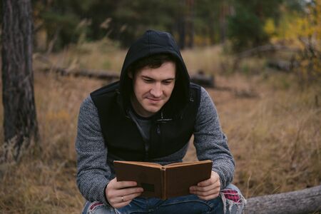 A young handsome man in casual clothes is sitting on a log and reading a book in the autumn forest.