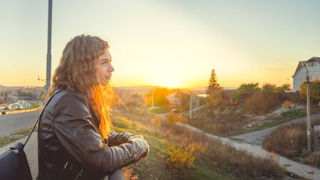 A young beautiful woman in a leather jacket with a black backpack stands thoughtfully on the bridge against the background of the urban district.の写真素材