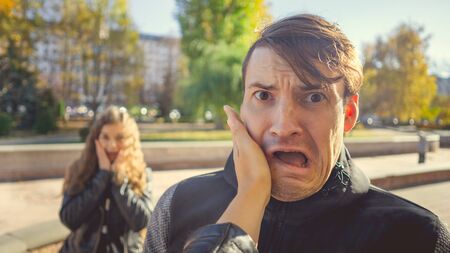 A womans hand hits a mans face in the autumn park. An emotional male is getting slapped in face, while shouting in a fear. A mistress is in a state of shock in the background.の写真素材