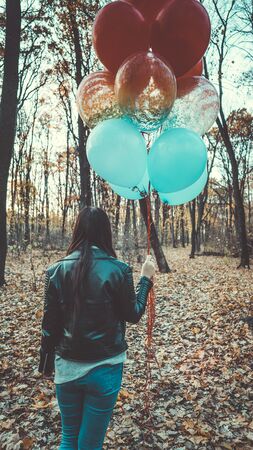 A young beautiful woman in casual clothes is holding the colorful balloons on walk in autumn forestの写真素材