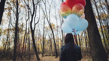 A young beautiful woman in casual clothes is holding the colorful balloons on walk in autumn forestの写真素材