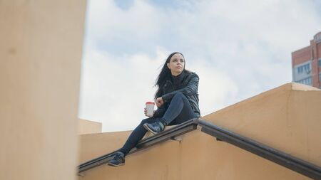 A young beautiful woman in casual clothes is sits on a concrete fence, looks away and holds a cup of coffee.の写真素材