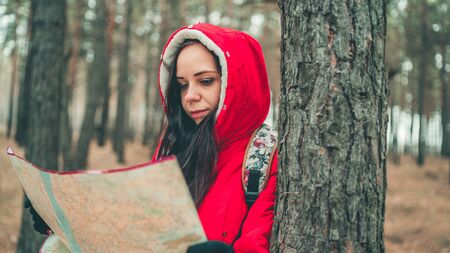 A traveling woman with map in woods. A portrait of the pretty woman with a backpack, standing near a tree in a cold weatherの写真素材