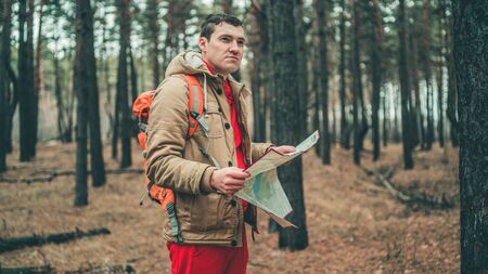 A traveling man with map in woods. A portrait of the handsome man with a backpack, standing near a tree in a cold weatherの写真素材