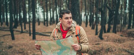A traveling man with map in woods. A portrait of the handsome man with a backpack, standing near a tree in a cold weatherの写真素材