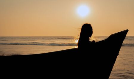Side view of dreamy lady standing near shabby boat on wet beach near waving sea and admiring cloudless sunset sky on resortの写真素材