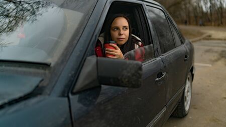 Long haired woman with disposal cup at front seat of car looking at window