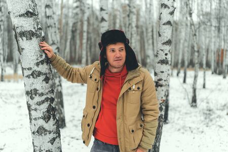 Serious young male wearing beige jacket over red hoodie and earflap hat standing on snowy forest and...