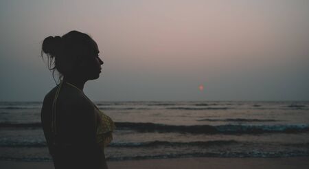Back view of unrecognizable female in swimwear standing on beach near waving sea in evening on...