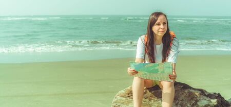 Barefoot woman with backpack sitting on driftwood and examining map while resting on sandy beach near waving seaの写真素材