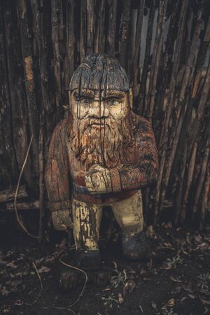 Wooden gnome near wooden fence on ground. Old cracked figure carved from wood. Garden gnome in...
