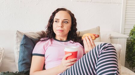 Female eats a juicy hot dog in a cozy bedroom. Concept: Temptation on a diet and fast foodの写真素材